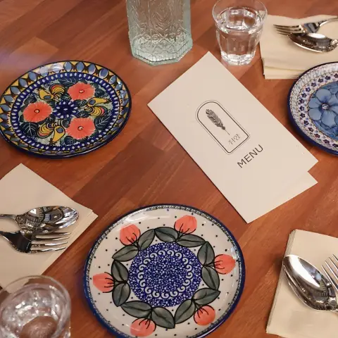 A wooden table set with three intricately patterned Polish pottery plates, silverware, glasses, and a closed menu card.
