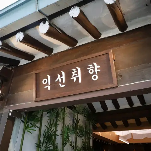 A close-up of the wooden signboard for "익선취향" (Ikseon-Chwihyang) above an entrance, framed by traditional hanok beams and decorative bamboo.