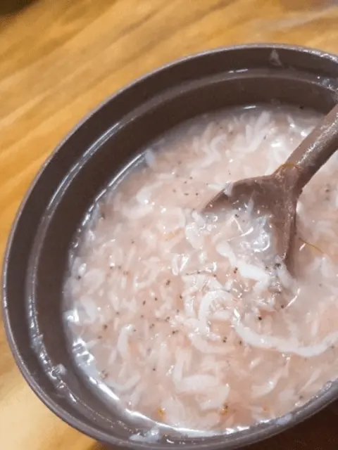 A close-up of a spoonful of Saeu-jeot, Korean salted baby shrimp, being lifted from a dark brown bowl. The bowl contains more salted shrimp and some liquid.