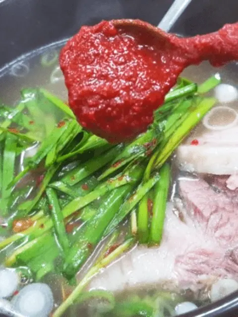 A spoonful of bright red sauce, likely gochujang or a similar chili paste, being added to a bowl of Korean soup containing sliced meat and green chives.