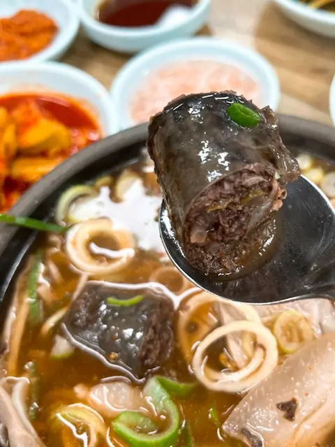 A spoonful of Sundae-gukbap, a Korean blood sausage and rice soup, being lifted from a dark bowl filled with broth, sliced sundae, and green onions. Various side dishes are visible in the blurred background.