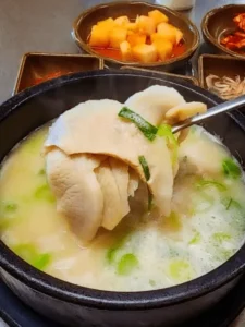 A spoonful of sliced pork and green onions being lifted from a steaming bowl of milky white Korean soup. Several small dishes of side dishes are visible in the background.