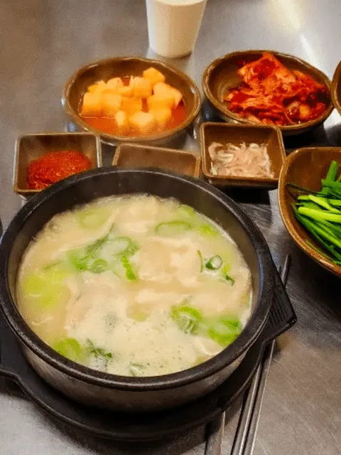 A top-down view of a table setting with a bowl of Gukbap, a Korean rice soup, surrounded by various small dishes of Korean side dishes (banchan) such as kimchi, pickled radish, and chives.