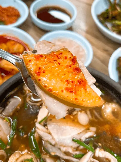 A spoonful of sliced meat topped with a bright orange, seasoned radish kimchi being lifted from a dark bowl of Korean soup containing meat and green onions. Several small dishes are visible in the blurred background.